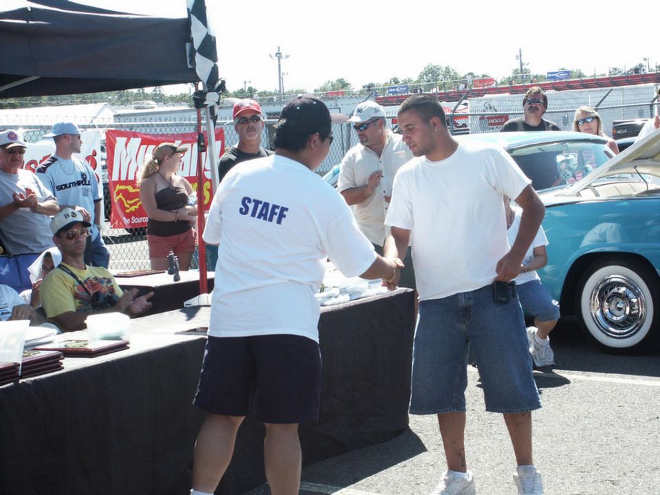 People at a car show, one in a STAFF shirt shakes hands; a turquoise 1956 Ford Fairlane is parked nearby
