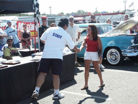 2001 Mercury Grand Marquis - A staff member shakes hands with a woman next to a turquoise 1955 sedan at a car show.