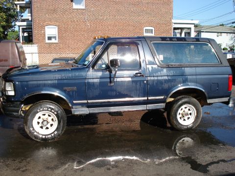 1994 Ford Bronco - A blue Ford Bronco with white wheels sits on a wet surface, reflecting the sky.