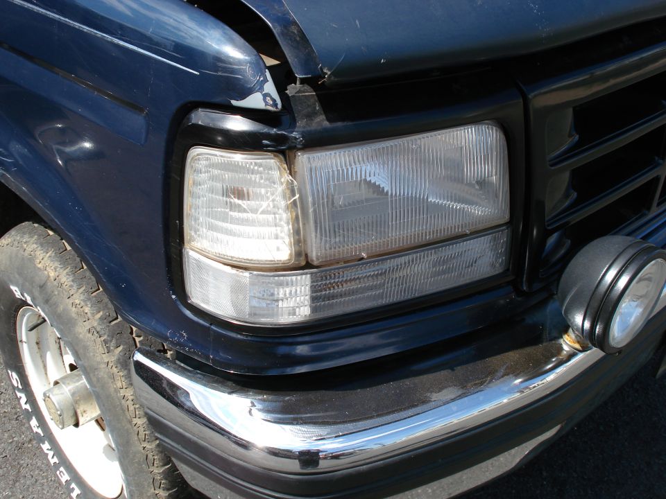 Dark blue Ford Bronco front corner with cracked headlight, chrome bumper, and tire. Hood is ajar