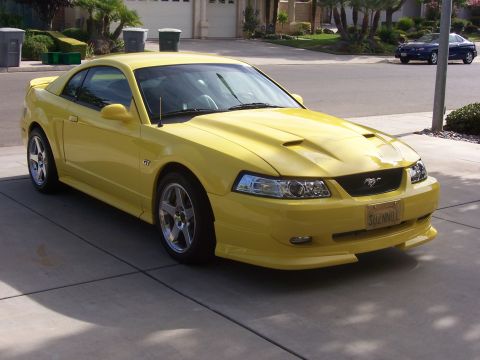 2002 Ford Mustang - Yellow 2002 Ford Mustang GT coupe parked on a driveway, showing its front 3/4 view with a hood scoop and GT badge.
