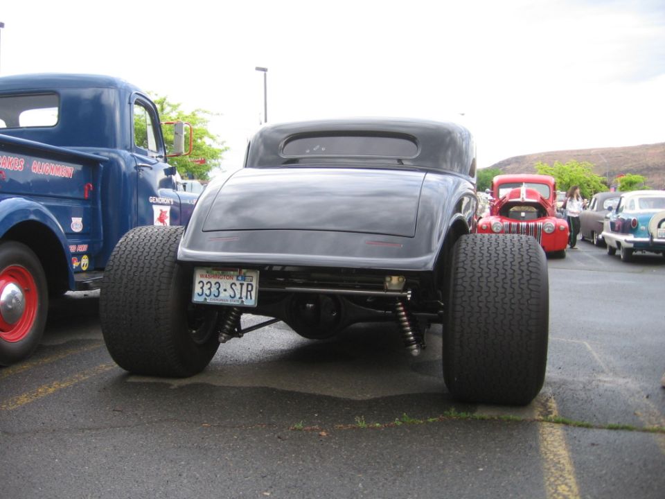 Rear view of a black hot rod coupe with wide tires. A blue truck is to the left, and a red car is behind it