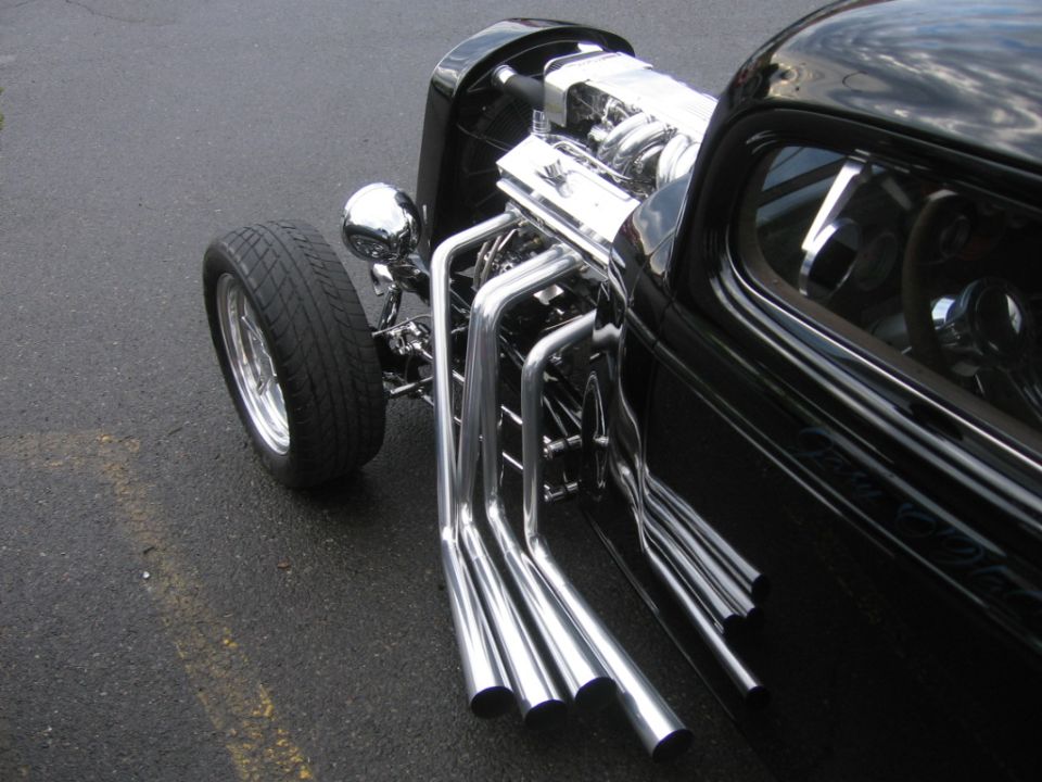 Black hot rod with chrome exhaust pipes and exposed engine, viewed from the front three-quarter angle