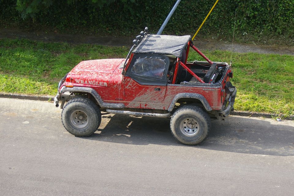 Muddy red Jeep Wrangler in profile with soft top folded down and tall whip antenna parked at a curb