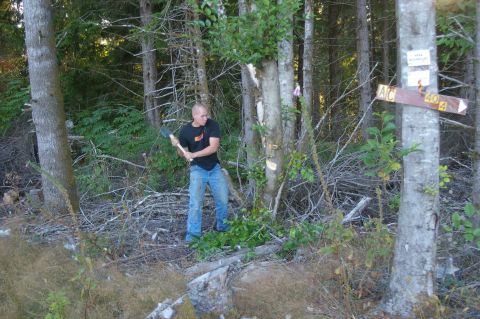 1998 Jeep TJ - Man chopping a tree in the woods with an axe. A trail marker sign is visible on a tree.