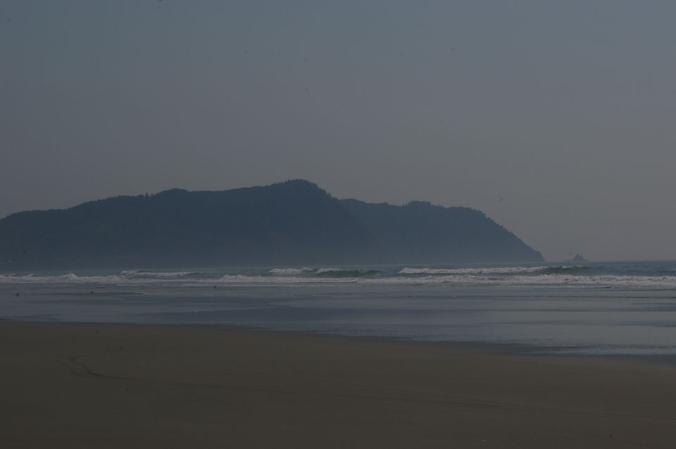 Beach with waves and distant hills under a hazy sky, with a website logo in the corner
