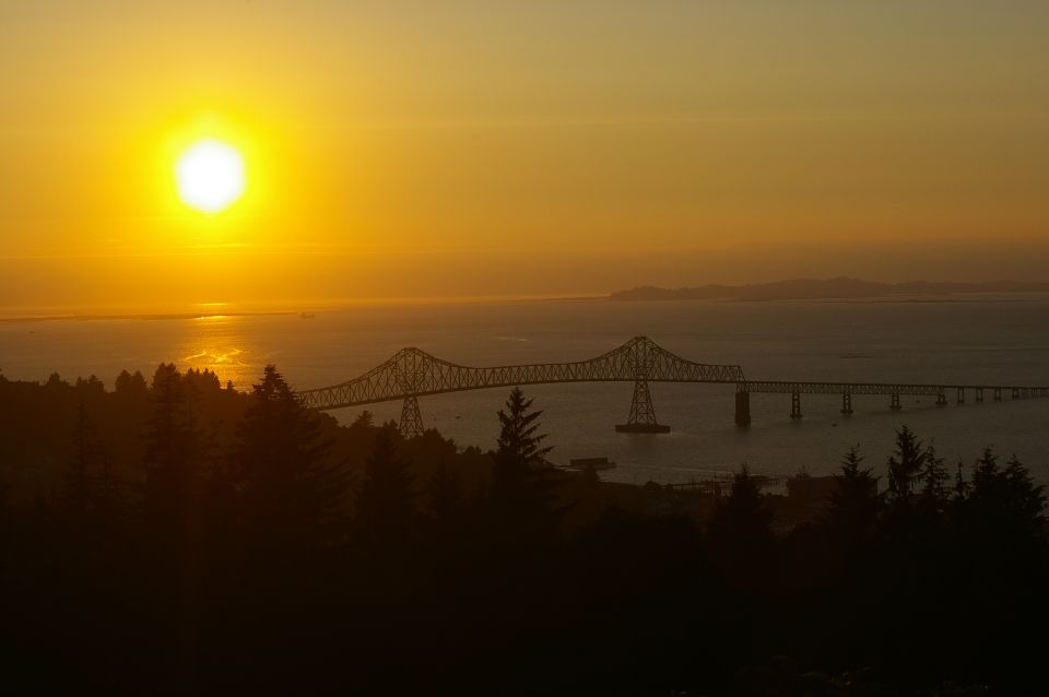 Sunset over a large bridge spanning a body of water, with trees in the foreground. Supermotors.net logo in the corner