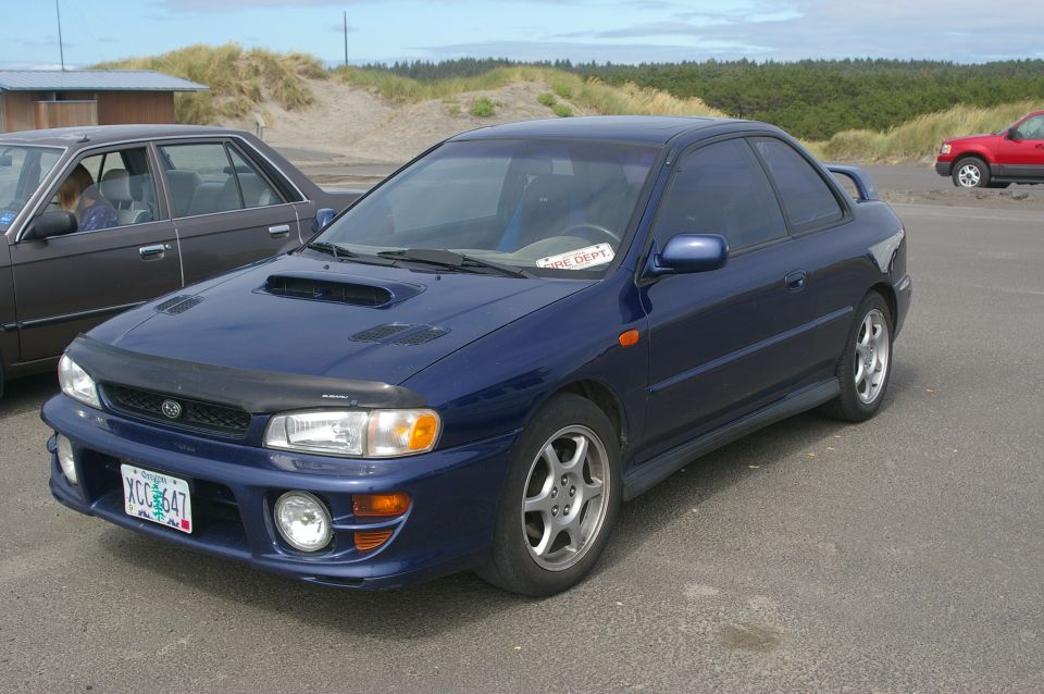 Blue Subaru Impreza coupe with hood scoop and spoiler, parked near dunes