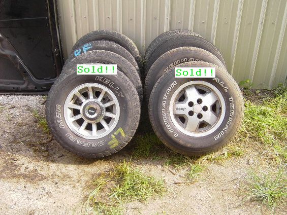 Two sets of Jeep wheels and tires, one with MSR Safari branding, the other with Jeep branding, stacked on dirt next to a black vehicle door