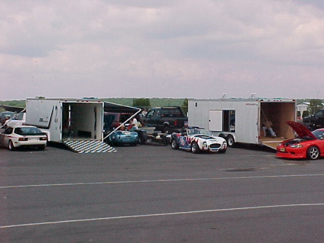 Race paddock with enclosed trailers, sports cars and a white Cobra replica