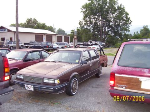 1979 Ford Bronco - A modified brown station wagon is parked in a lot with other vehicles, a prominent exhaust stack and roll cage.