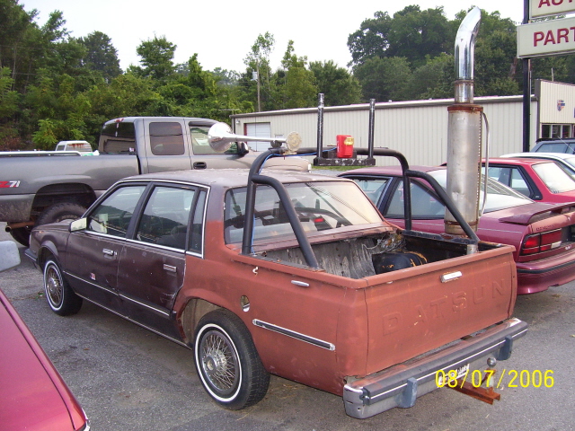 Custom Datsun pickup with roll cage and large exhaust stack parked next to a pickup truck and other cars