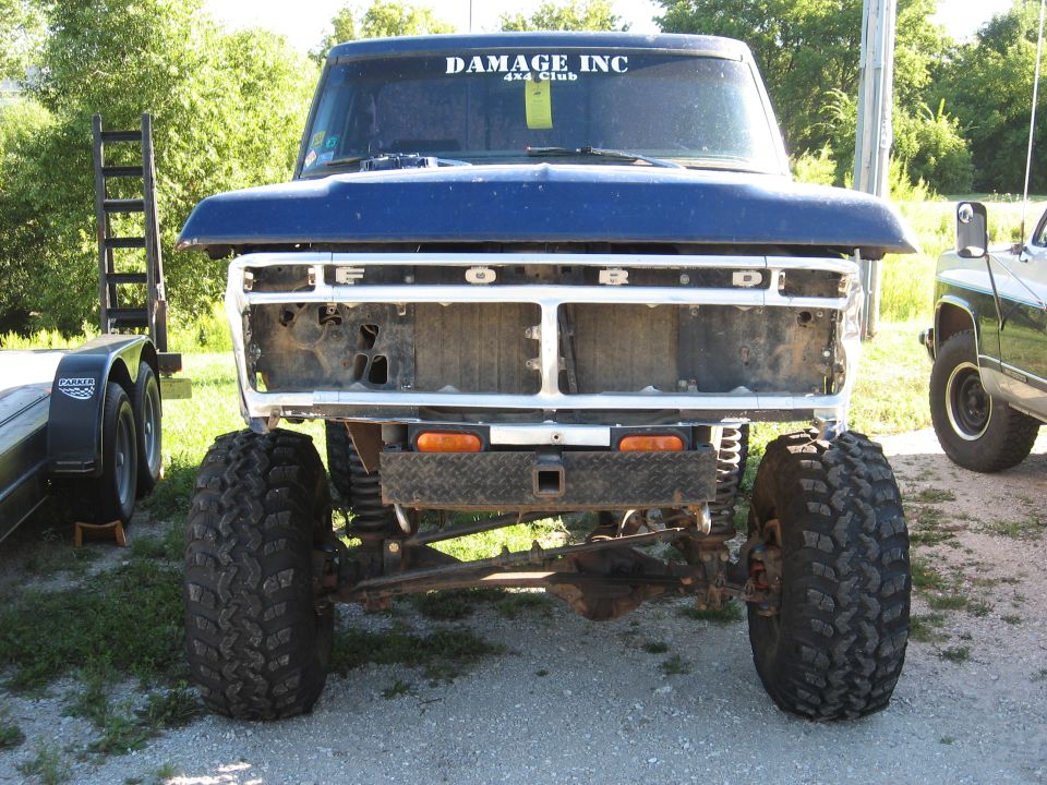 Blue 1977 Ford F-150 front view, missing grille, large tires, parked next to a trailer and another truck