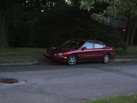 1998 Ford Taurus - A red 1998 Ford Taurus is parked curbside on a street with trees in the background.