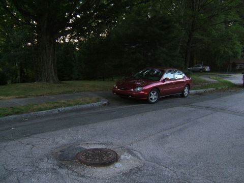 1998 Ford Taurus - A red sedan is parked on the side of a street, with a large tree and sidewalk to its left.