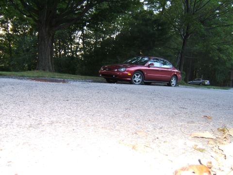 1998 Ford Taurus - A red sedan is parked on the side of a road with trees in the background.