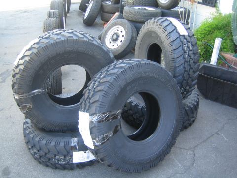 1990 Ford Bronco - Several new chunky mud-terrain off-road tires lean in a stack outdoors with more tires in the background.