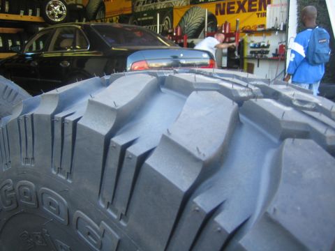 1990 Ford Bronco - Close-up of a large mud-terrain tire's aggressive tread block, with cars and a Nexen banner in the shop behind.