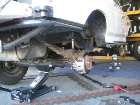 1990 Ford Bronco - underside of a lifted white truck on a floor jack, exposing the solid axle, leaf springs and brake rotor.