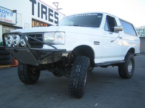 1990 Ford Bronco - lifted white Ford Bronco on mud-terrain tires with a tube bumper and roof lights parked outside a tire shop.