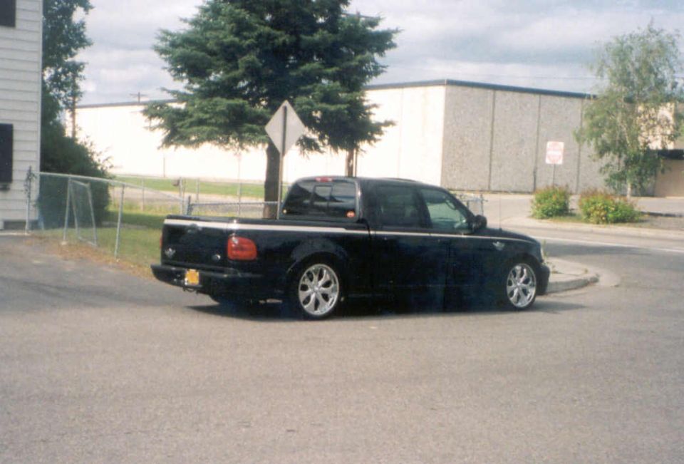 Black lowered Ford F-150 crew cab pickup on chrome wheels, rear three-quarter view in a lot