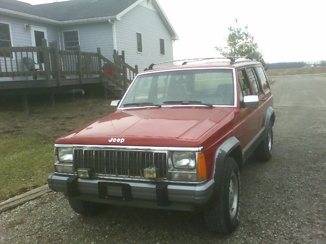 Red Jeep Cherokee parked on gravel, front 3/4 view. House and yard in background