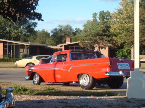 1971 Ford Ranchero - A bright red Ford Ranchero drives down a street lined with trees and mid-century modern houses.
