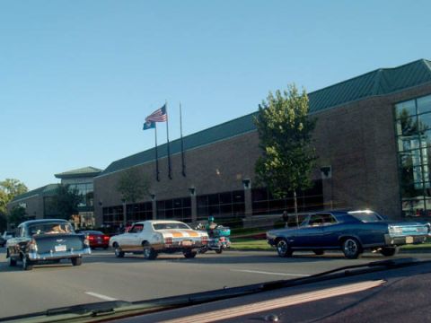 1971 Ford Ranchero - A blue 1967 sedan cruises past a building with American flags on a clear day.
