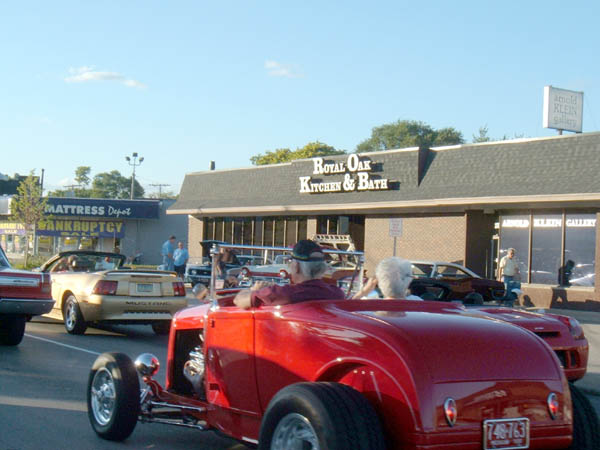 Red hot rod in foreground, gold convertible Mustang behind it, and other cars on street in front of businesses