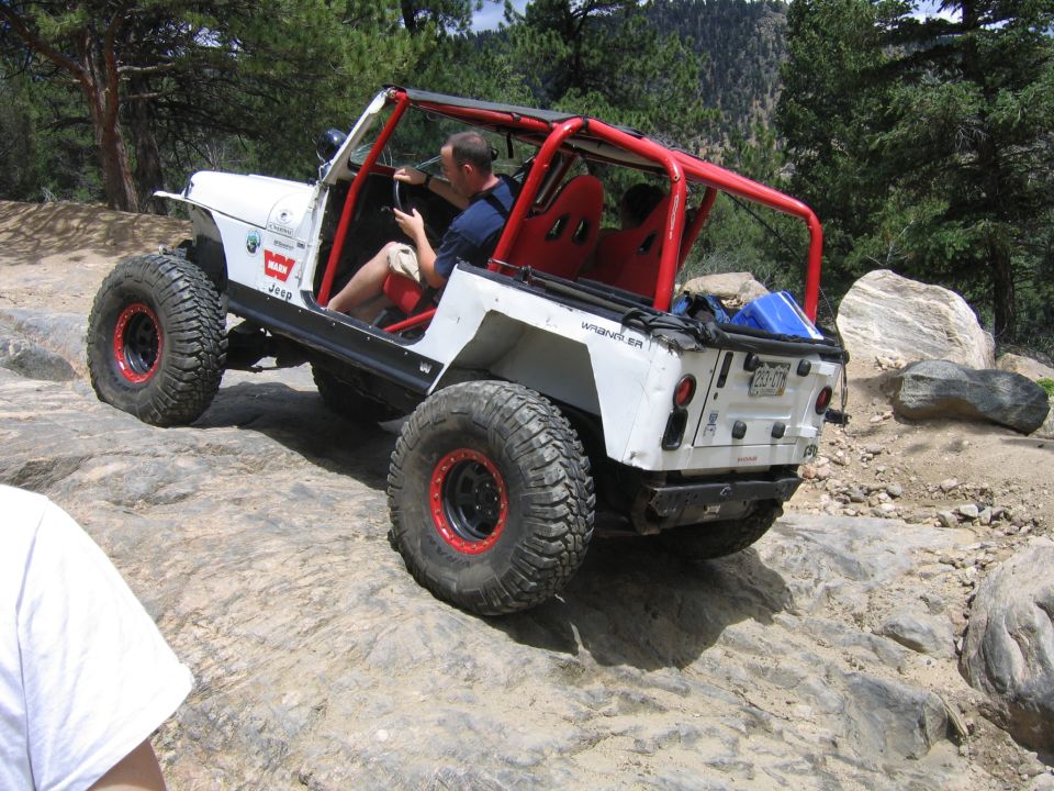 White Jeep Wrangler with red roll cage and seats, lifted on large tires, driving over rocks