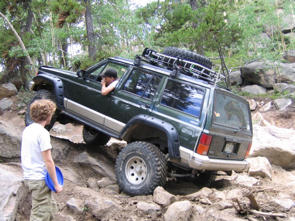 Dark green 1996 Jeep Cherokee with a roof rack and spare tire navigates a steep, rocky incline
