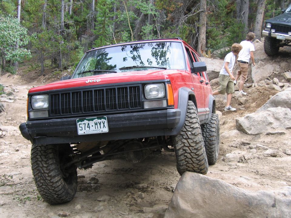 Red 1996 Jeep Cherokee navigating a rocky trail with two people walking behind it. Another green Jeep is visible further up the trail