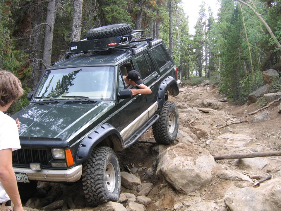 Dark green 1996 Jeep Cherokee with a roof rack and spare tire navigates a rocky off-road trail