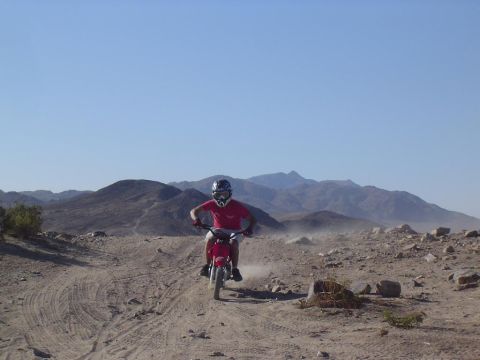 1985 Ford Bronco II - A rider on a red dirt bike navigates a dusty desert trail with mountains in the background.