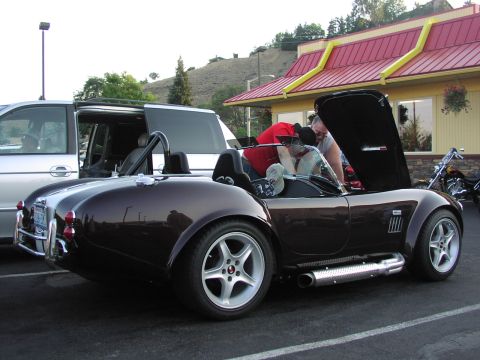 1979 Ford Bronco - A dark brown convertible sports car with its hood open is parked next to a silver minivan in a parking lot.