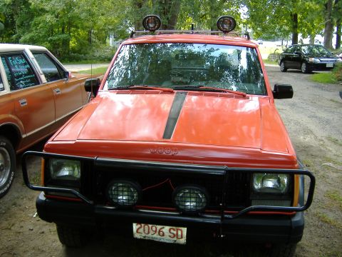 1990 Jeep Cherokee - An orange Jeep Cherokee is parked next to another vehicle, with a black grille guard and auxiliary lights on the roof.