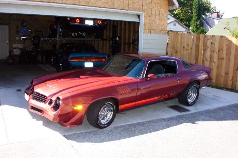 1979 Chevrolet Camaro - A red coupe with a distinctive side stripe is parked in front of an open garage, with another vehicle visible inside.