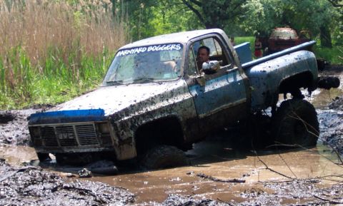 1978 Ford Bronco - Muddy GMC pickup truck stuck in a bog with "Barnyard Boggers" sticker on windshield.