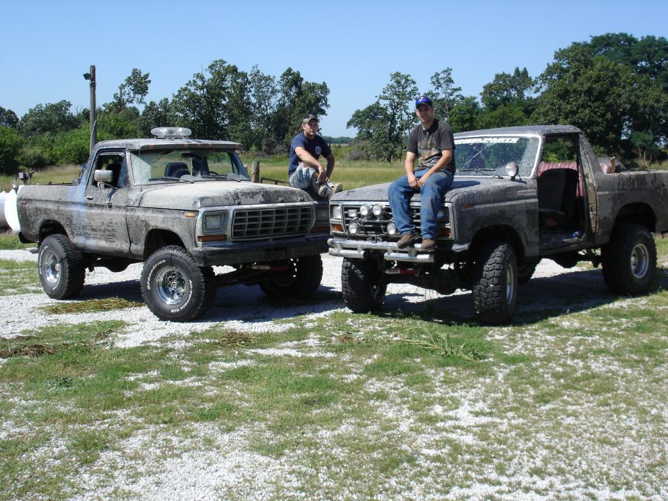 Two muddy Ford trucks parked outside, one with a light bar on top. Two guys are sitting on the hoods