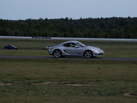 2001 Mercury Grand Marquis - A silver Porsche Cayman S drives on a track with 'POCONO' visible on a barrier in the background. Another car is seen in the distance.