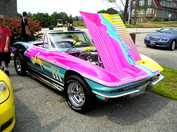1965 Chevrolet Corvette convertible with a vibrant pink, teal, and yellow graphic paint job, hood open, parked next to a yellow car