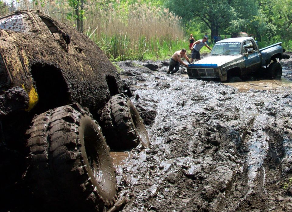 Two trucks stuck deep in mud, one on its side, with people trying to push the other