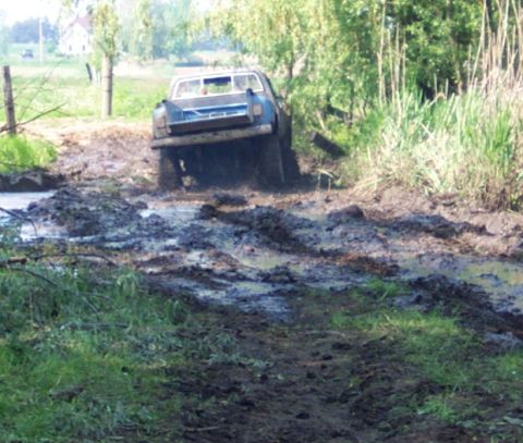 1978 Ford Bronco - A blue pickup truck navigates a challenging muddy path, surrounded by lush greenery.