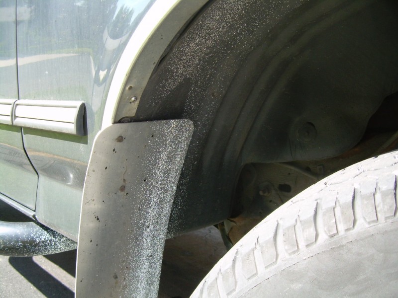 Close-up of a vehicle's rear wheel well with sand and a mud flap. Tire is visible