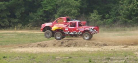 1979 Ford F-150 - Two monster trucks catch air while racing on a dirt track, kicking up dust.