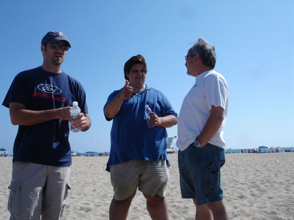 Three men stand on a sandy beach under a clear blue sky. The man on the left wears a dark blue t-shirt with a Ford logo