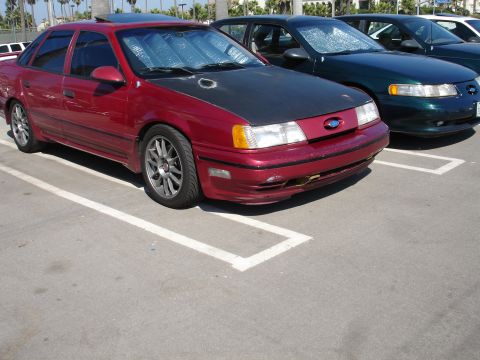 1995 Ford Taurus - A red 1995 Ford Taurus sedan with a black hood and aftermarket wheels is parked in a lot next to other vehicles.