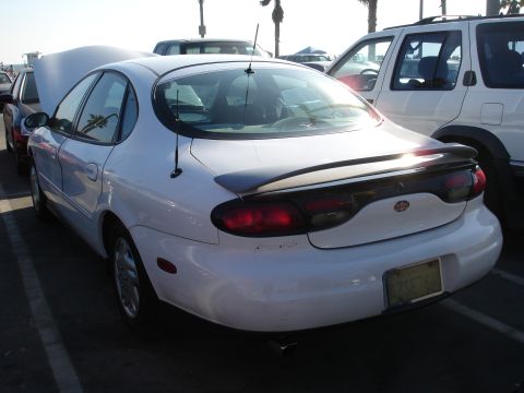 1995 Ford Taurus - A white sedan is parked in a lot with its hood open, alongside other vehicles.