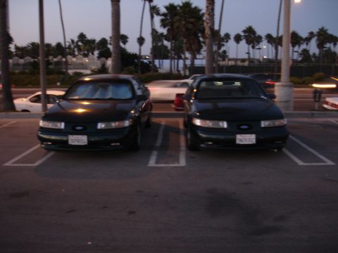 1995 Ford Taurus - Two dark 1995 Ford Tauruses are parked side-by-side in a parking lot, with their front ends facing the camera.