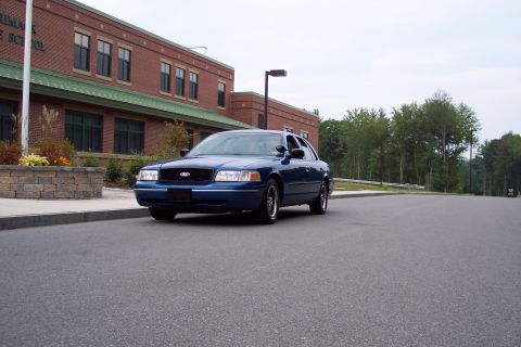 2001 Ford Crown Victoria - A blue 2001 Ford Crown Victoria is parked on the side of the road in front of a school building.